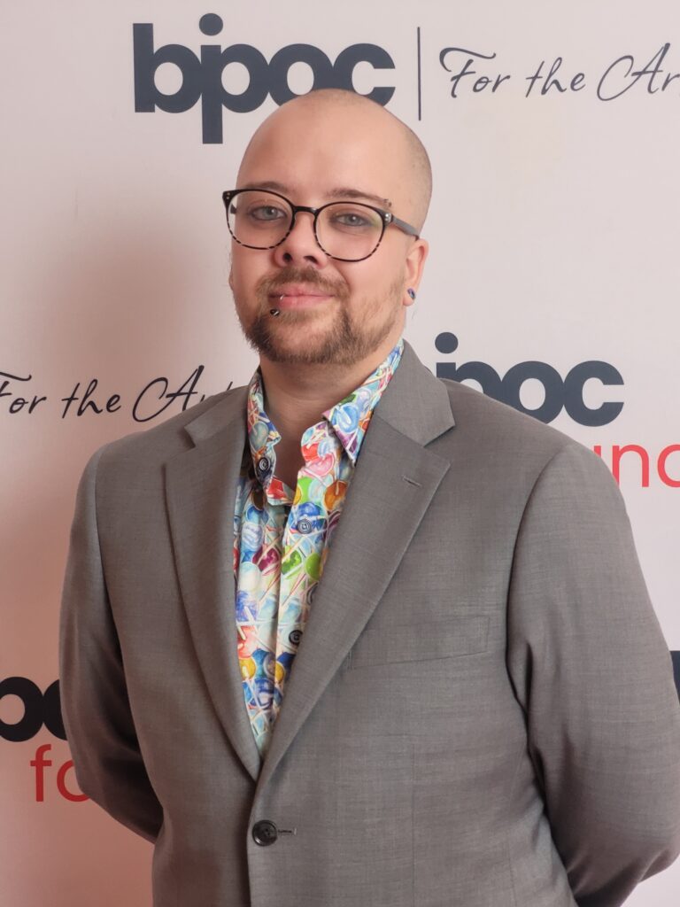 Man with shaved head in grey sport jacket and colourful shirt.